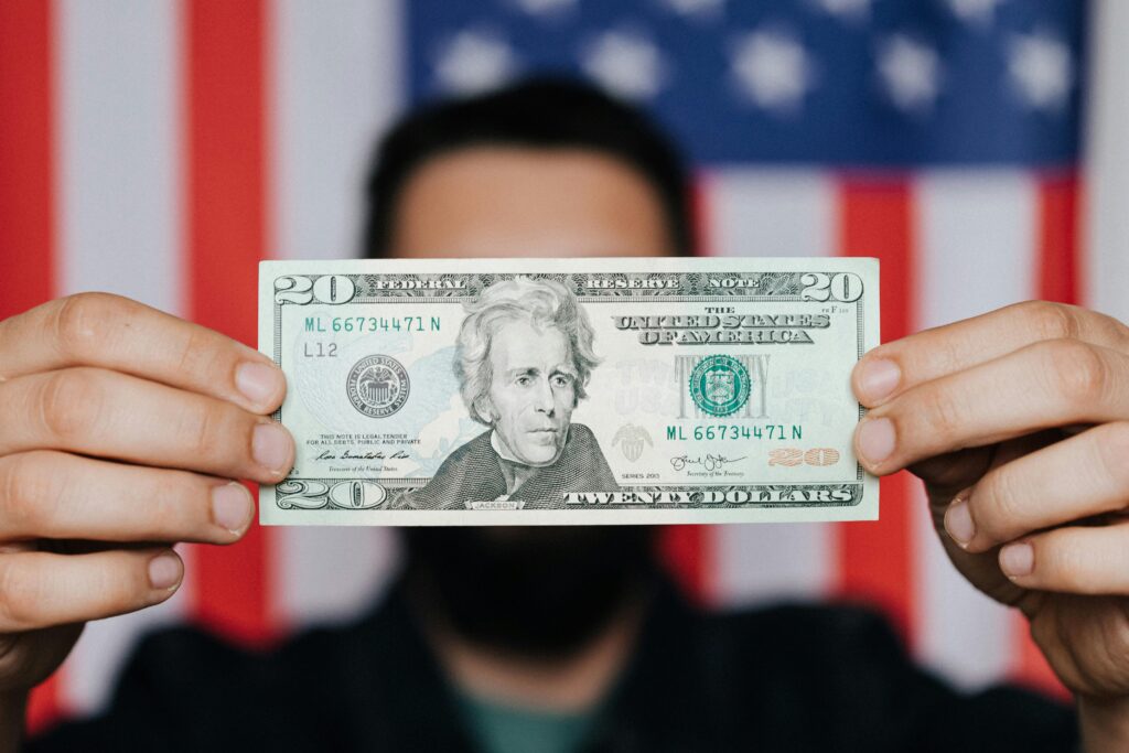 a peson shows a US currency bill infront of his face the us flag is in background